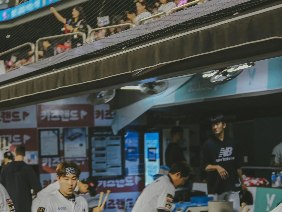 KBO players near the dugout with fans pressed against the netting above, representative of the crowd energy at a Korean ballpark
